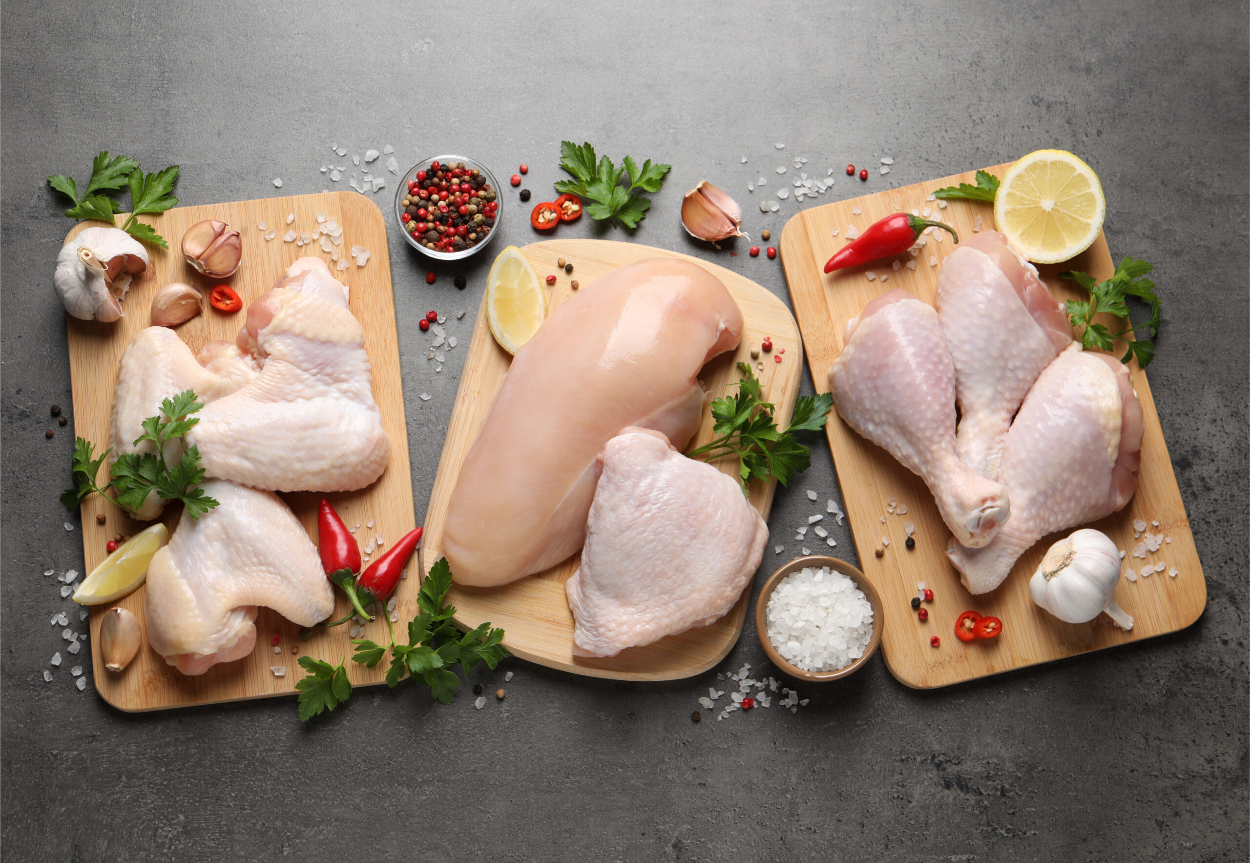 Fresh raw chicken pieces displayed on wooden cutting boards, including chicken breasts, thighs, and drumsticks, garnished with garlic, herbs, lemon, and chili peppers, set against a textured gray background. Ideal for meal preparation and cooking inspiration.