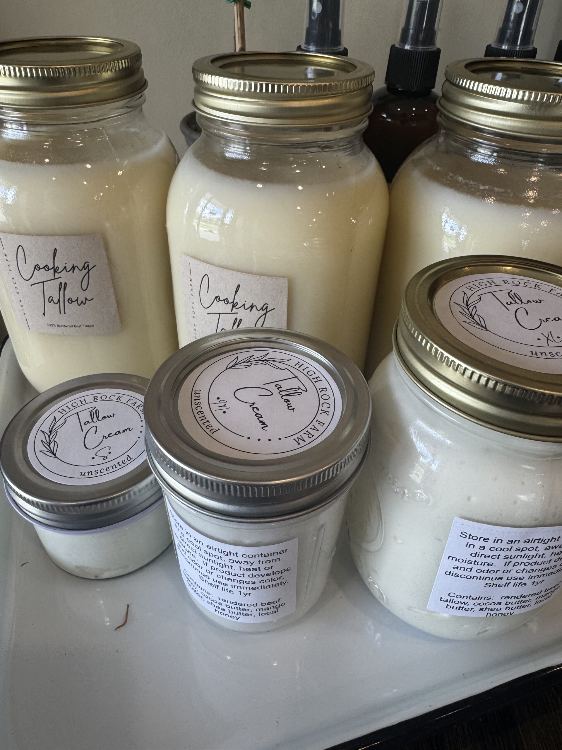 Jars of cooking tallow and tallow cream displayed on a shelf, featuring labels with product information and storage instructions. The jars vary in size and design, showcasing unscented and natural ingredients for cooking and skincare.