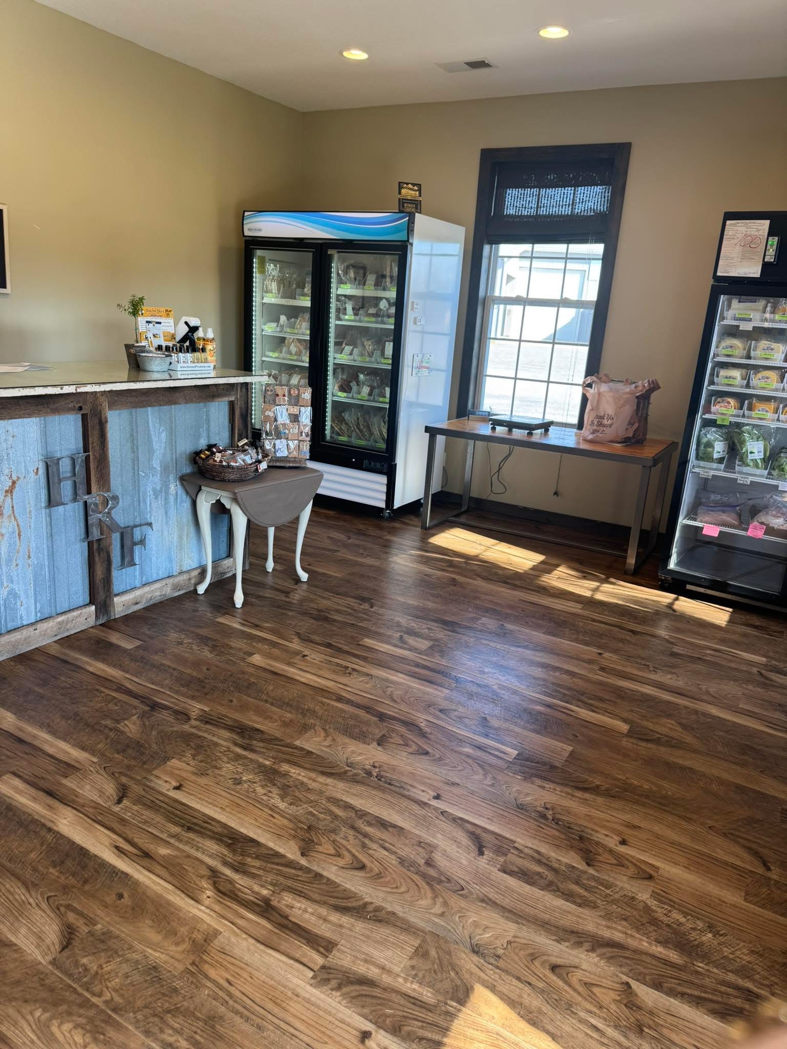 Interior view of a cozy retail space featuring polished wooden floors, a rustic counter, and two glass display refrigerators stocked with various products. A small table near the window holds a bag and promotional materials, creating an inviting atmosphere for customers.