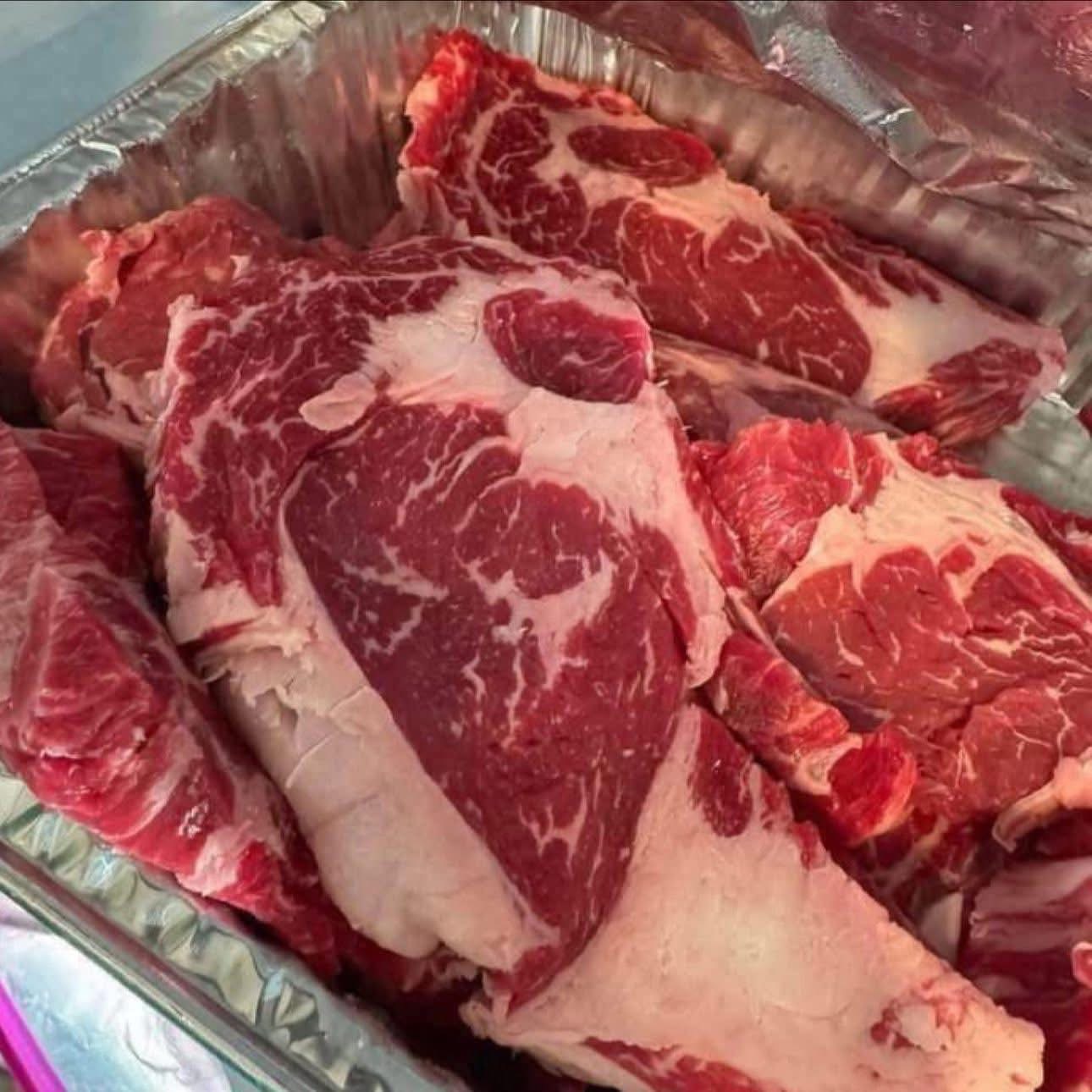 Assorted cuts of raw beef, including marbled ribeye and chuck, displayed in a metal tray. The meat features a rich red color with visible marbling, indicating tenderness and flavor.