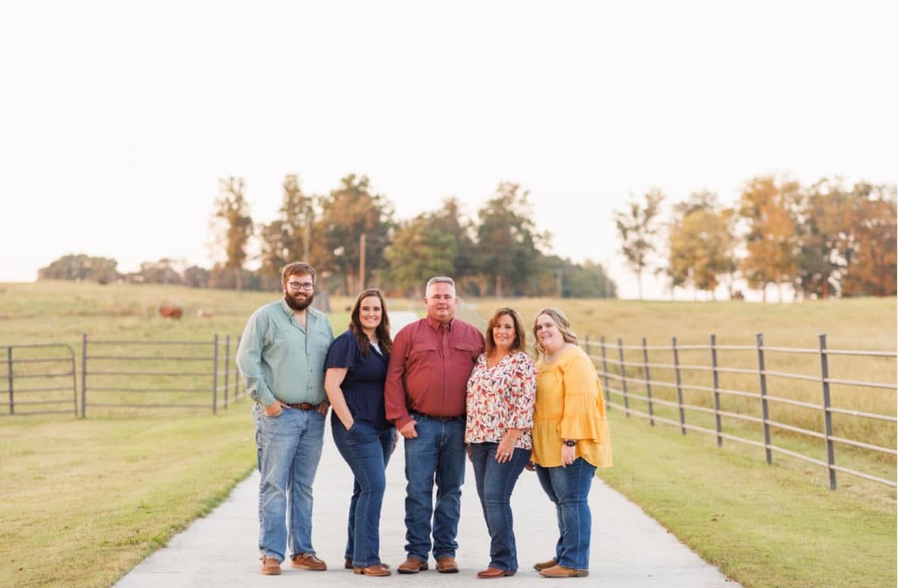 Kayla - IMG_9371 Family portrait on a rural path, featuring five adults dressed in casual attire with a scenic landscape of fields and trees in the background. The image captures a warm, autumn atmosphere.