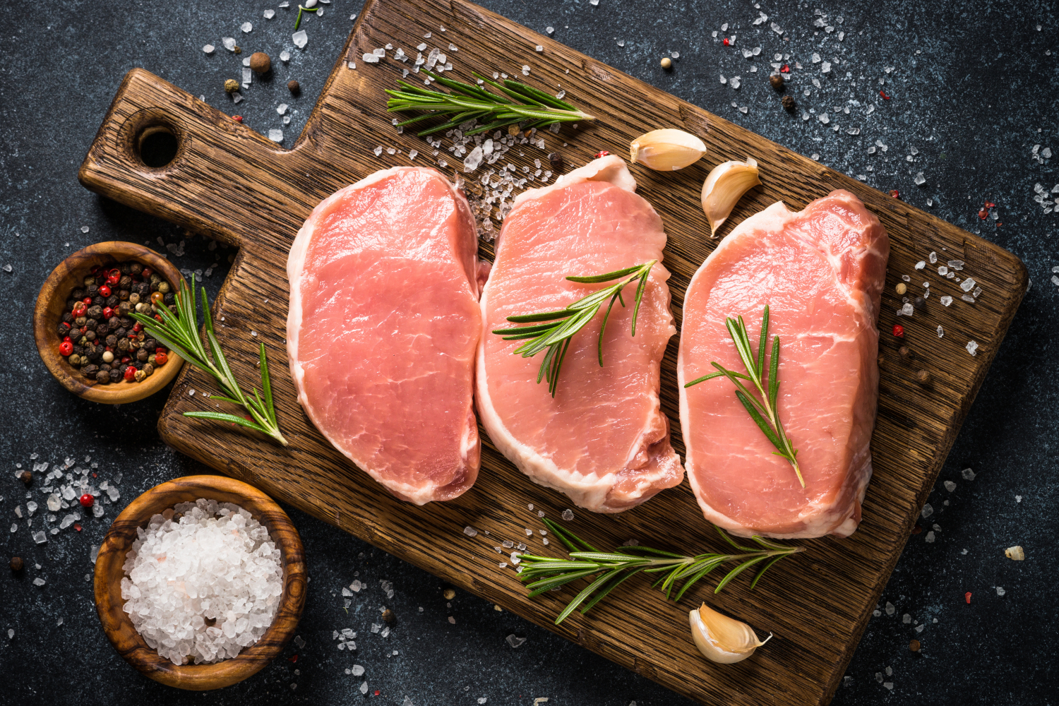 Fresh pork chops garnished with rosemary and garlic on a wooden cutting board, surrounded by coarse sea salt and colorful peppercorns. Ideal for recipe blogs and cooking websites.