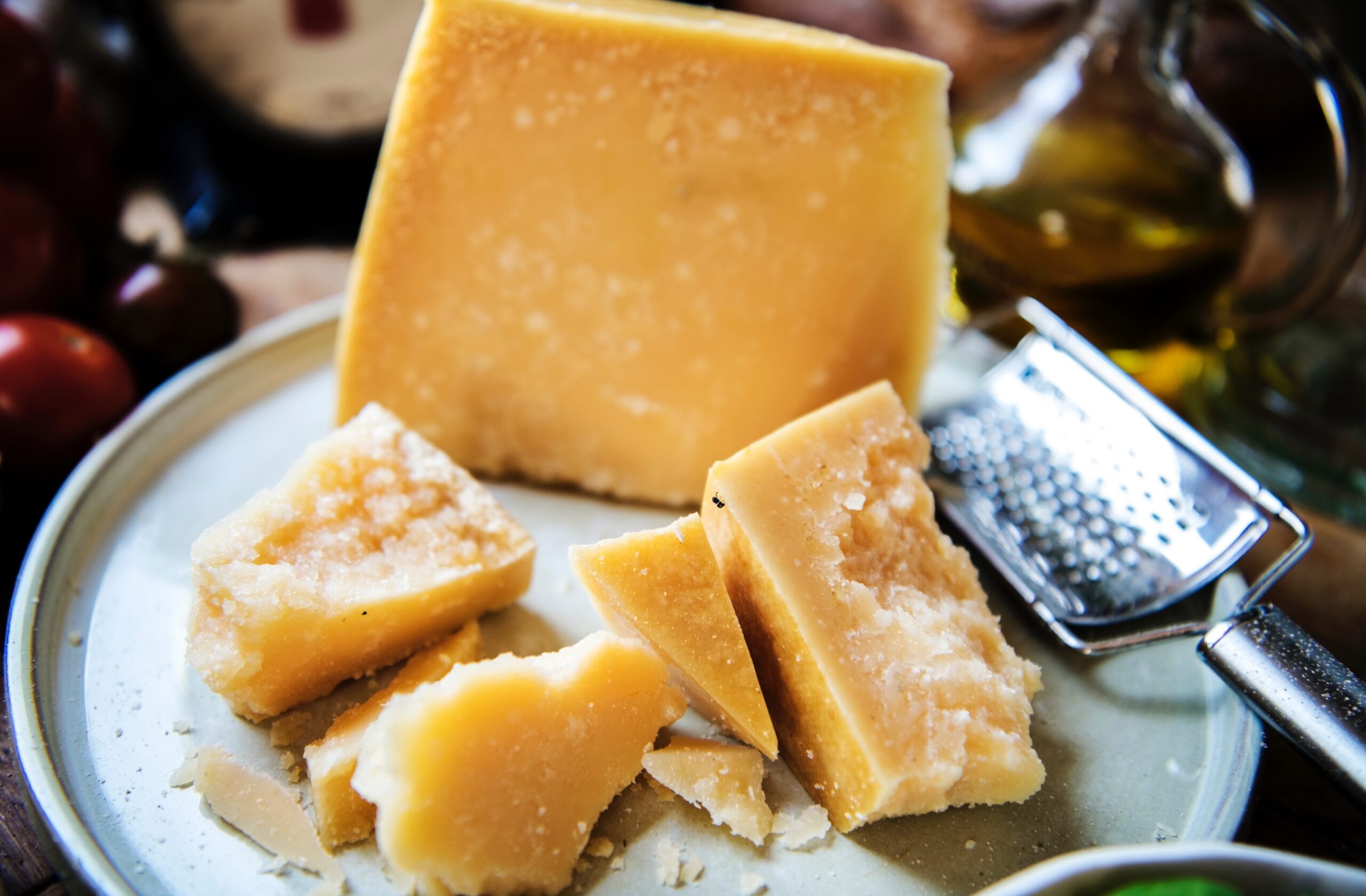 A plate with pieces of aged cheese, including a large block and smaller chunks, accompanied by a cheese grater and a drizzle of olive oil in the background. The setting features fresh tomatoes, highlighting a rustic culinary theme.
