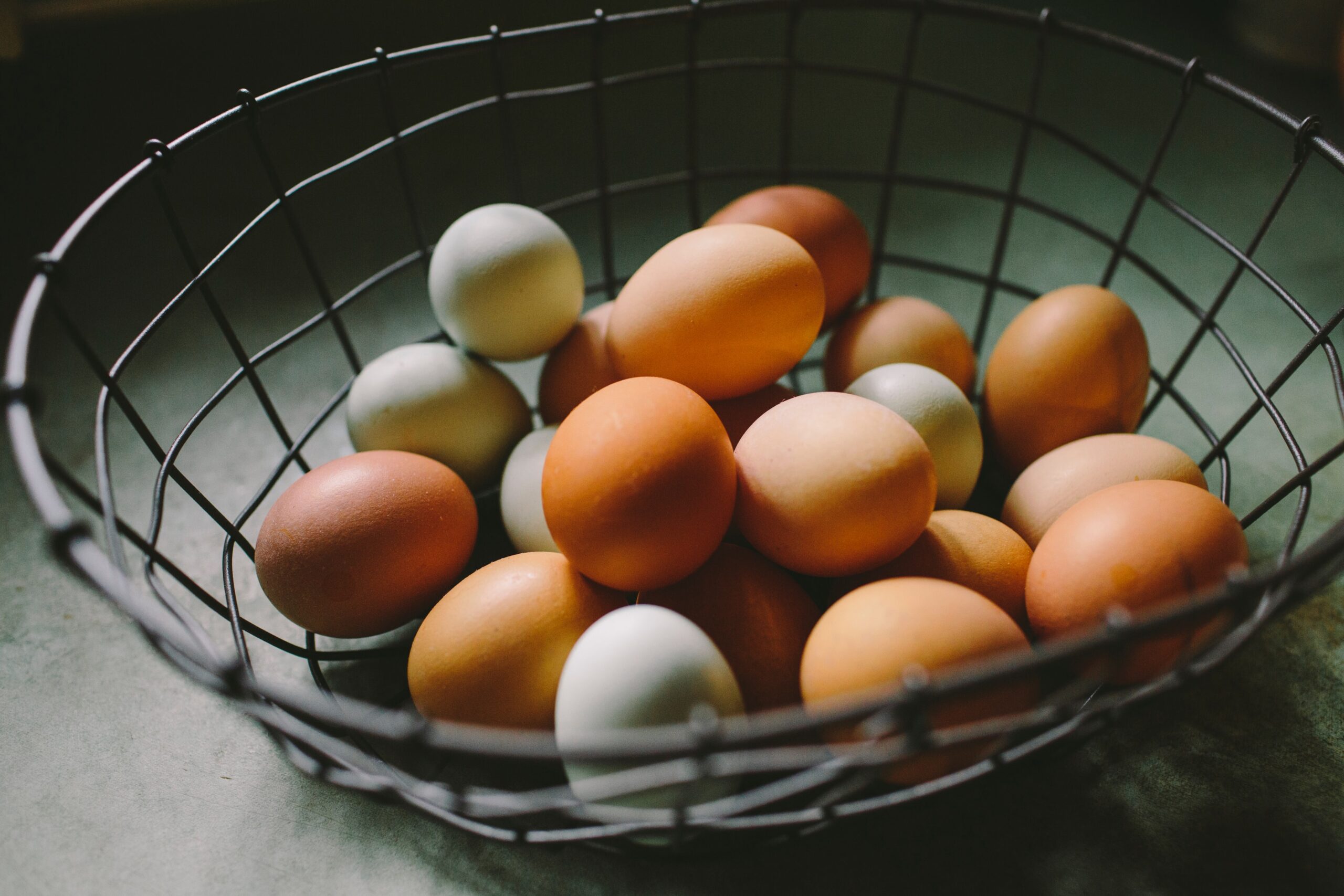 A wire basket filled with a variety of eggs in different colors, including brown and pale green, resting on a textured surface.