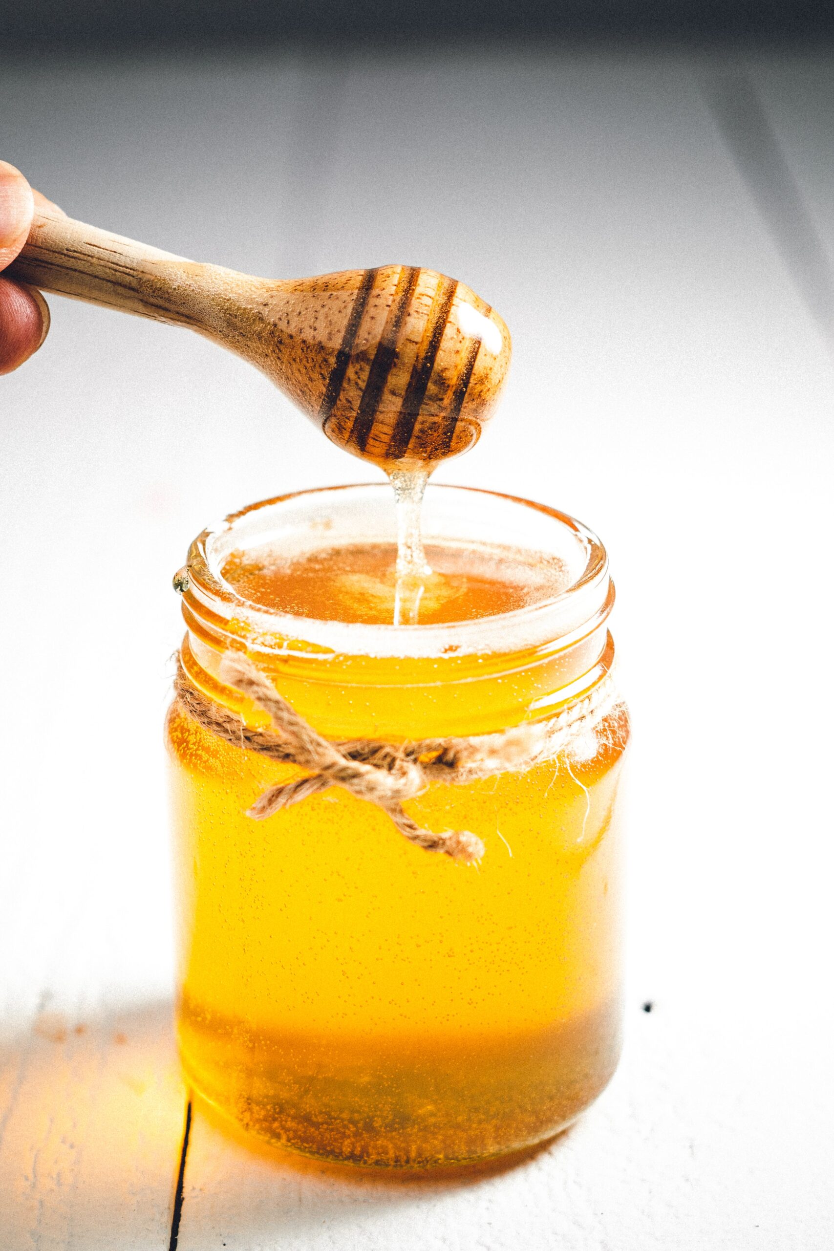 Hand pouring honey from a wooden dipper into a glass jar filled with golden honey, showcasing its rich texture and color against a bright background.